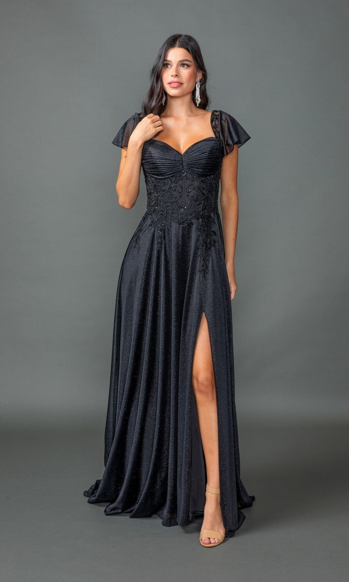 A woman wears the Dancing Queen 4496 Short-Sleeve Long Formal Dress in black, featuring glitter lace, a sweetheart neckline, flutter sleeves, and a thigh-high slit, standing against a plain gray background.